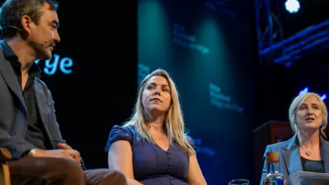 Sarah sits between Tim Wu on the left and Carole Cadwalladr on the right on stage at hay. she is looking towards tim. she has shoulder length blonde hair and is wearing a blue short sleeve dress. Carol is blond and in a suit jacket and Tim is turn towards Carole and Sarah in profile with short dark hair and a dark suit. 