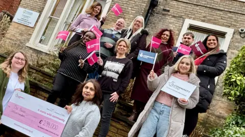 Nine women and two men stand outside a building holding pink and blue lottery flags with two woman at the front holding a cheque for £294,000. The main part of the group are stood on stairs at different levels.