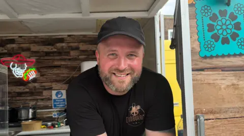 Carl Condon is wearing a black t shirt with his company logo and a black baseball cap. He is smiling and leaning over the counter of his food van from the inside. A neon sign with Mexican paraphernalia is visible on the van's wall behind him