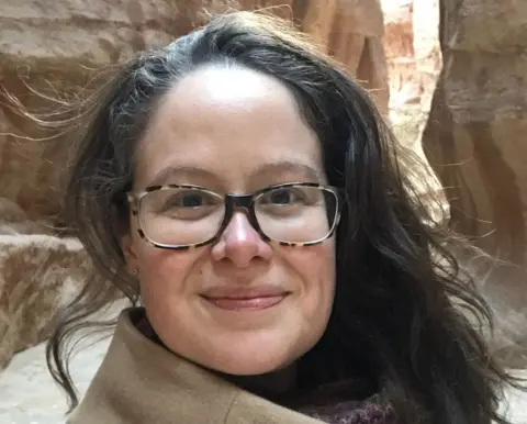 Dr Kirstin Anderson, a dark haired woman wearing glasses photographed in front of a rocky background