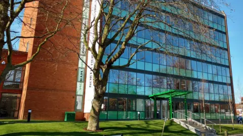 The front of North Hertfordshire Council's building in Letchworth Garden City. There are two trees without leaves in front of the building, the building itself is six stories high and has a lot of windows with a set of steps leading towards its entrance. The sky is blue behind the building.