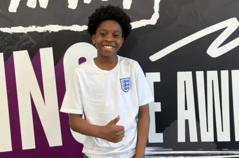 BBC A boy in an England football shirt gives a thumbs up to the camera.