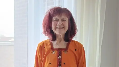 Marie Curie Hazel, 65, has a red bobbed haircut and is wearing an orange kaftan-style top. She is in her living room and looking directly into the camera.