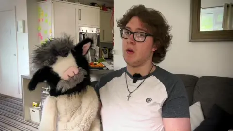 A young boy with dark hair wearing a crucifix necklace and a white and grey t-shirt. He's holding up a fluffy cow puppet. He's in a house and there's a grey sofa and the edge of a kitchen behind him.