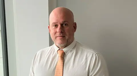 Det Chief Insp Neil Fuller. He is bald with a white stubble and brown eyes. He is standing against a white wall next to a window. He is wearing a white shirt and peach tie.