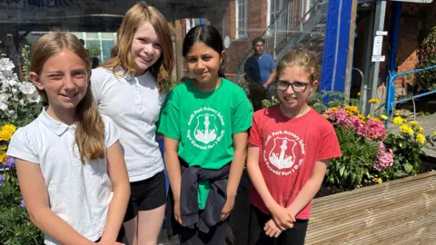 The four girls are pictured on their school yard in front of flower beds. They are all smiling looking at the camera. Seren, on the left, is wearing a white t-shirt and has long blonde hair, which is draping on her left shoulder. Next to her is Eleanor, who again is wearing a white t shirt, black shorts and has light brown shoulder-length hair. Fatima is standing next to Eleanor. She is wearing a green Roath Park Primary School t-shirt and has a blue jumper tied around her waist. Fatima has black hair in a ponytail, with a middle parting. Ada, on the right, is wearing a red Roath Park Primary School t-shirt. She wears glasses and has light brown hair which is in a ponytail.