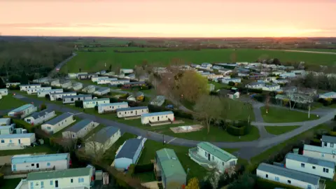 An aerial shot of the Cakes and Ale Holiday Park. Its shows a large plot of land with caravans and buildings on it. There is a sunset in the distance. 