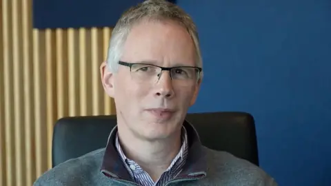 Mark Norman/BBC Pfizer scientist Dr Nicholas Kitchin looks at the camera with a blue wall behind him.