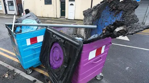  REX/Shutterstock Debris fills charred wheelie bins on County Road