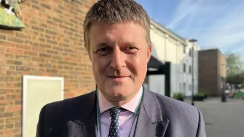 BBC Richard Clewer wearing a grey suit and pink shirt standing in front of a building on a sunny day and looking at the camera