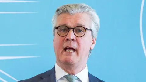 Malcolm Offord is mid-speech. He has short white hair, and wears black round glasses and a grey suit with a pale green tie. He stands in front of a sky blue background