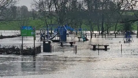 Jonathan Sterritt A child's play-park which is flooded with water. A number of trees without foliage can be see around the park. Green fields can be seen in the background. 