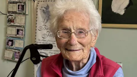 Penny Lees 107-year-old Mollie Silver smiles while sitting down inside her home. Pictures and artwork are on the wall behind her. She is wearing a blue turtle neck jumper and a red gilet.