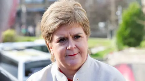 A woman with light-coloured hair photographed outside the Scottish Parliament in a close-up shot. She is wearing a white jacket and is visible from the shoulders up. 