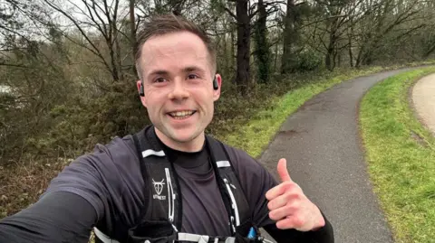 Man gives thumbs up on a path with grass at the side. He is in running gear.