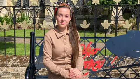 Alex Bulat sitting on a bench wearing a beige dress