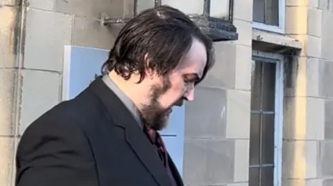 David Polwarth leaving court. He has medium-length black hair and beard. He is wearing a black jacket over a grey shirt and burgundy tie. He is looking down as he is snapped leaving the building.