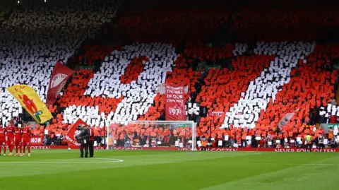 Fans in a packed stand at Anfield stadium display the number 97 against a red backdrop while players stand for a minute's silence on the pitch