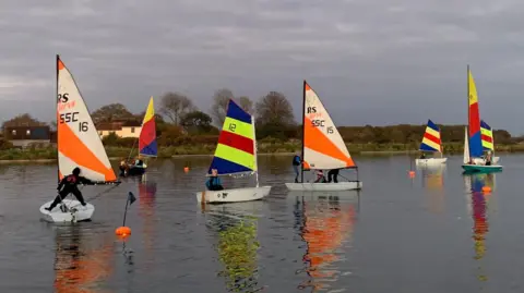 Sailing boats on the lake, sails flag colours are red and cream as well as blue, yellow and red.