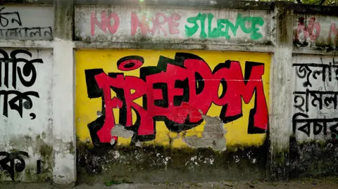 Mural painted on a wall reads "FREEDOM" in bright red letters, on a yellow background. Above that text are the words "No more silence".