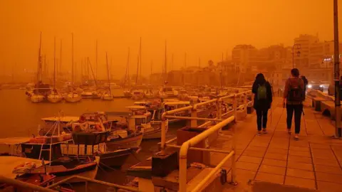 Stefanos Rapanis/Anadolu via Getty Images People walk along a waterfront on Crete with boats, buildings and the sea in the background. The sky is orange. Photo: 1 April 2026