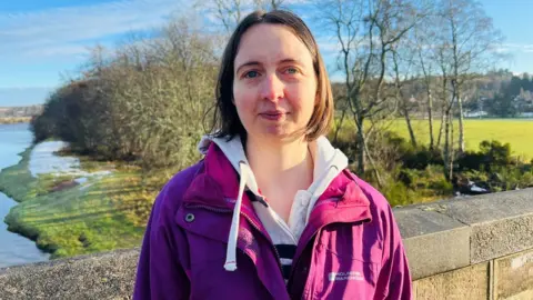 Charlotte Davys standing on a bridge and looking at the camera, with a river and trees in the background.