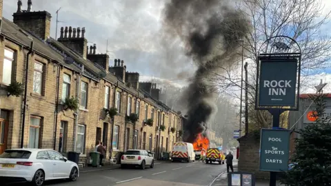 Daron Healy A white van is on fire in a street of terraced houses. There are intense orange flames and a large plume of black smoke rising into the sky. A few people are standing outside houses watching.