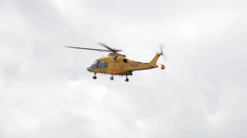 Grimsby News & Pictures Agency A yellow air ambulance flies away from the scene of the crash. The helicopter is set against a bright but cloudy sky.