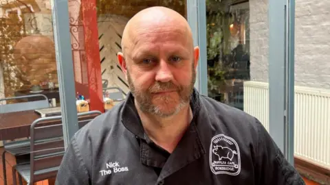 Charlie Stubbs / BBC There is a man who is bald. He has a brown beard with some grey hairs in it. He is wearing a black chef uniform with 'Nick The Boss' printed on it's right side and 'Truffles Cafe' printed on the right.