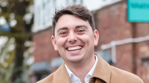 Matter Adam Root is wearing a beige jacket over a white shirt. He is standing in front of a brick building and smiling. He has short dark hair.