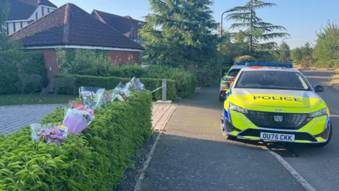 Holly Nichols/BBC The scene at Carnoustie Drive in Great Denham. On the left is a hedge which has plastic-wrapped flowers sticking into it, and beyond is an entrance to a driveway with police tape across it. On the right are two police cars parked partly on the pavement. Beyond it is another detached house and trees.