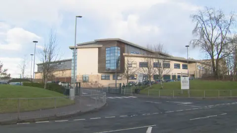 A view from across the street of Kirkintilloch High School - a large sign with the school name on it is sitting on grass, while a road leads into the premises. The building is mostly tan and brown shades, with a car park nearby. 