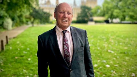 Jim Allister in a pink shirt and patterned floral tie, green tweed blazer, smiling stood on grass with Westminster in the background