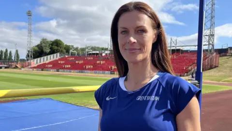 ANDY WATSON/BBC Emily MacDonald, who has long, brown hair and is wearing a blue Great Britain athletics shirt, is standing on the track with the grass pitch behind her.