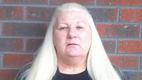 Scottish Prison Service Rona Mackenzie standing in front of a brick wall. She is looking directly at the samera and has long white hair.