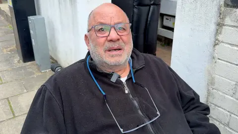 Yehia Hassan, who is wearing glasses and is in a wheelchair outside his house, has an anguished expression  