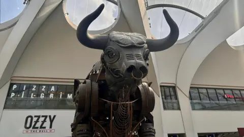 A photo of a mechanical bull in the middle of a train station, it has a flat cap on and is grey, matching the colour of it's head.
