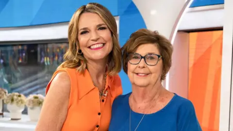 Savannah Guthrie, in a bright orange sleeveless top and patterened orange hued skirt, smiles beside her mother Nancy, in dark-rimmed glasses and a deep blue T-shirt, on a TV news set