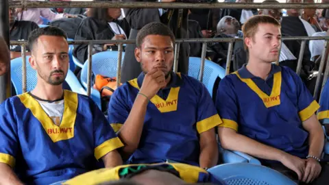 (L-R) Benjamin Zalman- Polun, Marcel Malanga and Tyler Thompson in blue and yellow prison uniforms wait to hear the verdict in their trial on 12/09/2024.