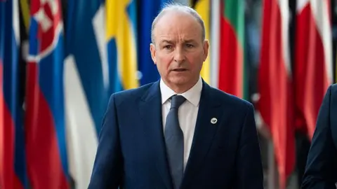 Getty Images A man in a navy suit wants in front of a number of international flags. He is wearing a white shirt and navy tie.