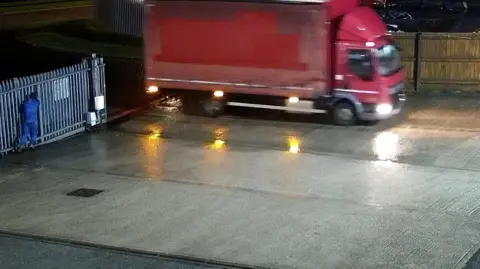 A still image provided by Blancomet shows a red HGV driving onto the firm's concrete forecourt through metal security gates. A man in a blue outfit stands next to the gates. Text on the CCTV image give the date as 18/01/2026, a time of 22:15:01 and says Front Entrance.
