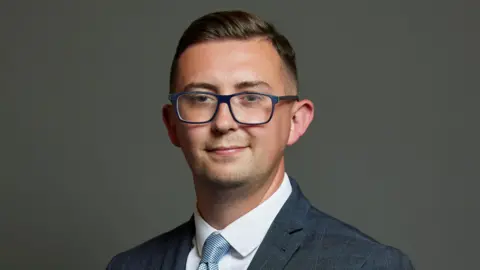 Connor Naismith, the Labour MP for Crewe and Nantwich, wears blue-rimmed glasses and has dark hair, parted on his left side. He has stubble on his chin. He wears a dark blue suit with slight light-blue checks on it with a white shirt and blue tie.