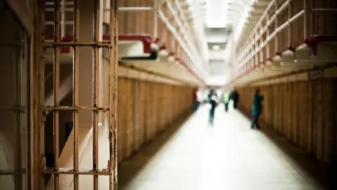 Image shows the corridor of a prison with cells visible 