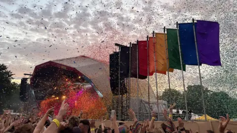 A large stage erected on the Downs in Bristol. There are rainbow coloured flags to the right of the stage. Crowds have their arms in the air as rainbow confetti falls from the sky.