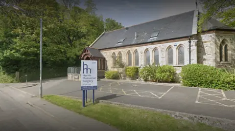 The image shows the outside of the Harris Memorial Surgery. The building is made from stone and has arched windows. There is a white sign in front of the building with the name of the surgery on in blue writing. There is a car park in front of the building with marked spaces. There is lots of  greenery, trees and bushes, around the building.