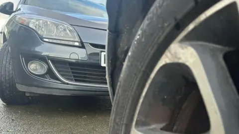 A close up of a car tyre, with the bumper of a grey car parked on wet concrete behind it.