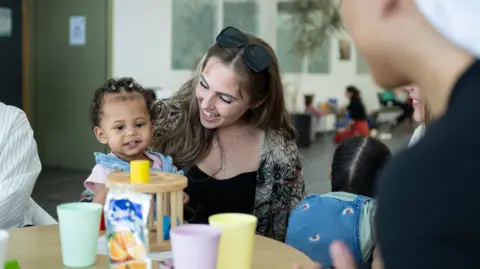Christy Nunn Zara sitting at a low table with her toddler daughter on her lap, they are both smiling and looking at a wooden toy on the surface. There are several plastic cups on the table and other parents and children in the large room behind