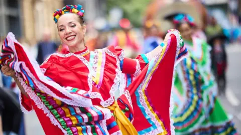 A woman with her brown hair pinned back holding up her traditional dress which is predominantly red with white lace and green, pink, yellow and blue ribbon during a parade. She wears large gold dangle earrings. 