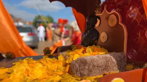 Jon Wright/BBC A small bronze statue of a Hindu king sits on a wooden carrier with orange petals