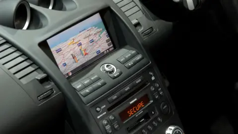A car dashboard has lots of buttons and controls in a black material. There is a screen in the centre of the console with a sat nav map.